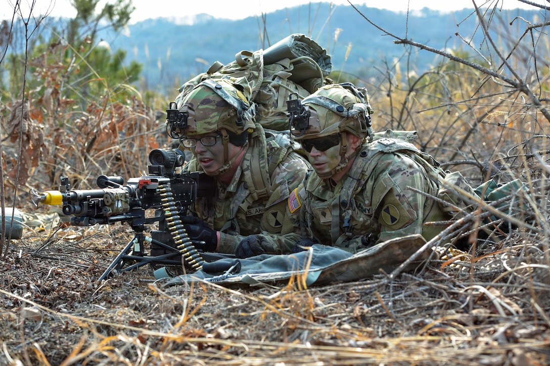 Two soldiers in camouflage lie next to each other in a field as one aims weapon loaded with ammo.