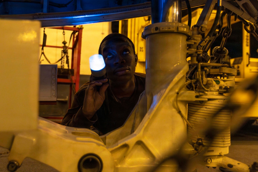 A kneeling sailor uses a flashlight to look at a partially visible aircraft in the dark.