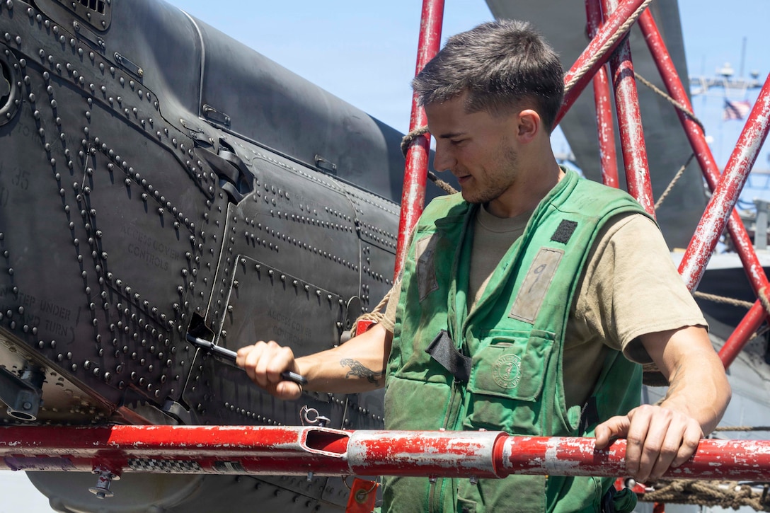 A sailor in a green vest on a red lift works on black partially visible aircraft aboard a ship during the day.