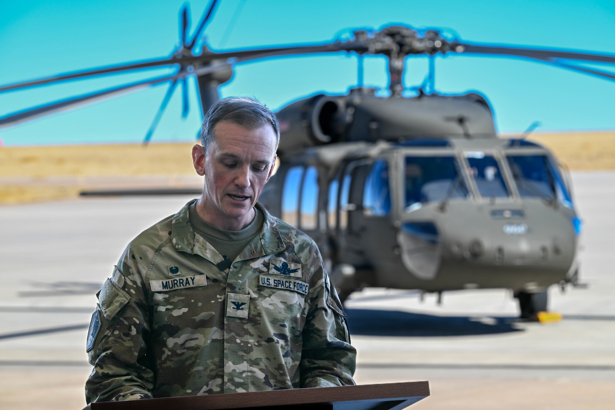 man speaks at podium in front of helicopter