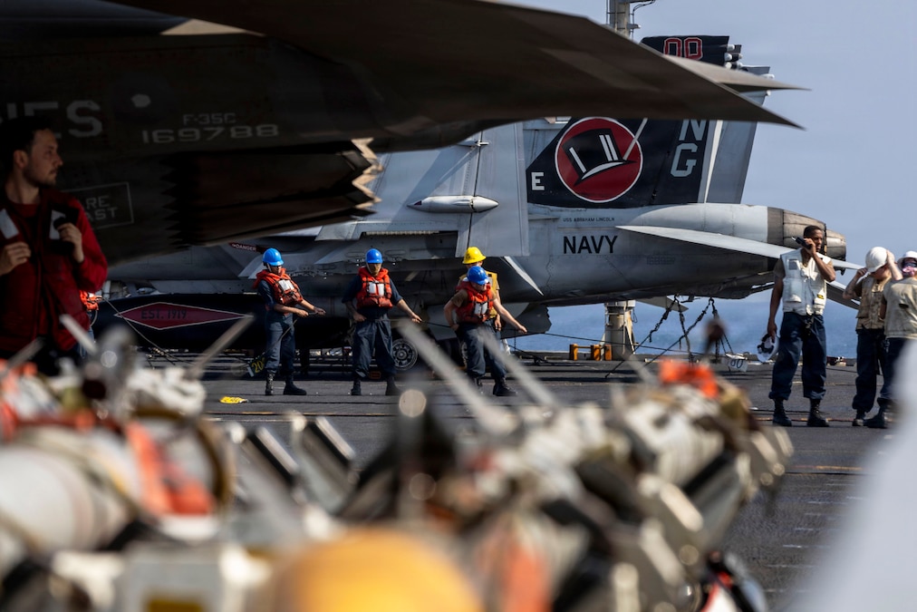 A group of sailors to the left pull a line while a group in white vests gather in a circle near parked aircraft aboard a ship at sea.