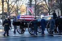 A U.S. flag-draped casket is being transported on a horse-drawn caisson down a street with military member marching alongside.