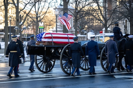 A U.S. flag-draped casket is being transported on a horse-drawn caisson down a street with military member marching alongside.