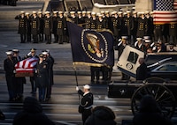 Military service members in ceremonial uniforms are carrying a U.S. flag-draped casket on the left, there is a hearse and caisson on the right, and a Sailor is holding the U.S. Presidential flag in the middle while a large group of Navy cadets in dark coats are seen in the background saluting.