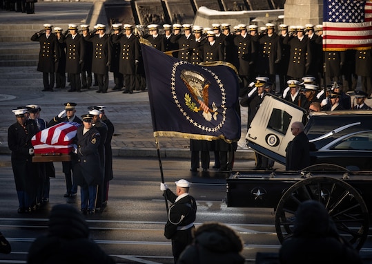 Military service members in ceremonial uniforms are carrying a U.S. flag-draped casket on the left, there is a hearse and caisson on the right, and a Sailor is holding the U.S. Presidential flag in the middle while a large group of Navy cadets in dark coats are seen in the background saluting.