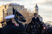 An Army Soldier in ceremonial uniform is riding a horse that is near other horse, which are pulling a caisson down a street, while someone salutes in the foreground and a flag is seen with buildings visible in the background.