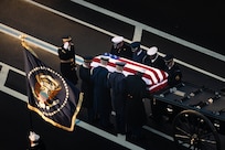 Service members from each branch of the military dressed in ceremonial uniforms are seen from above, lifting a US flag-draped casket off of a caisson, with one member standing a saluting and another holding the flag that represents the official colors for the U.S. president.