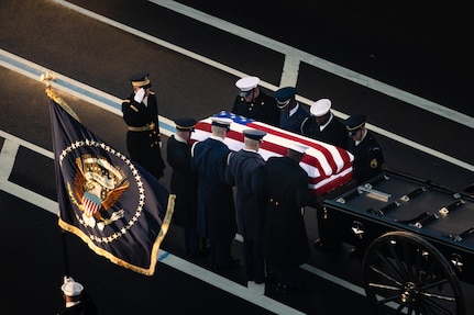 Service members from each branch of the military dressed in ceremonial uniforms are seen from above, lifting a US flag-draped casket off of a caisson, with one member standing a saluting and another holding the flag that represents the official colors for the U.S. president.