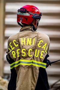 An Army soldier dressed in a beige firefighter uniform and red helmet is turned away from the camera and has the words Technical Rescue in yellow on the back of the jacket.