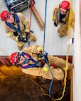 An Army soldier dressed in a beige firefighter uniform and red helmet is rappelling up a wall, with others looking up from below.