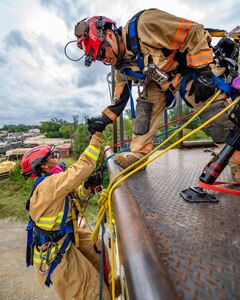An Army soldier dressed in a beige firefighter uniform and red helmet is being pulled up onto the top of a tower by a similarly dressed soldier after rappelling up the tower.