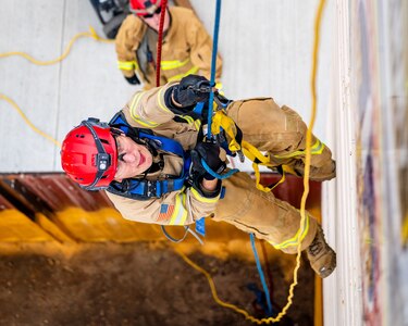 An Army soldier dressed in a beige firefighter uniform and red helmet is rappelling down a wall, with others looking up from below.