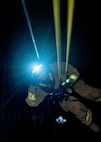 A Soldier dressed in a beige firefighter uniform with yellow reflective tape and a flashing light on his helmet is rappelling down a dark shaft.