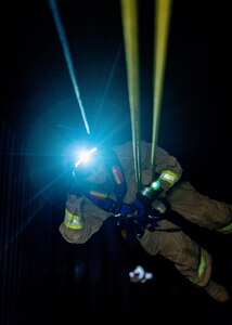 A Soldier dressed in a beige firefighter uniform with yellow reflective tape and a flashing light on his helmet is rappelling down a dark shaft.