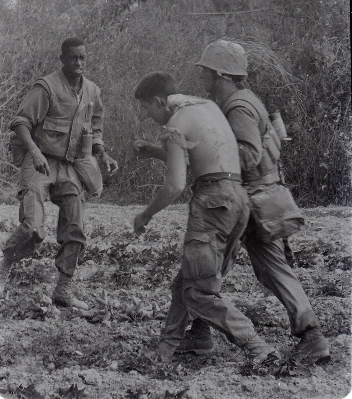 A man wearing a military uniform has his arm slung under a shirtless man to help him walk through a grassy area. Another man, dressed in a military uniform, walks in their direction.