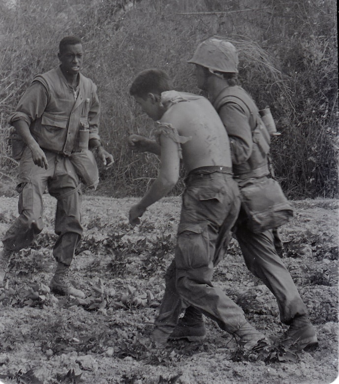 A man wearing a military uniform has his arm slung under a shirtless man to help him walk through a grassy area. Another man, dressed in a military uniform, walks in their direction.