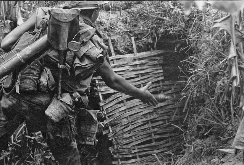 A man wearing several pieces of combat gear slung over his back throws a grenade into a thatched hole.