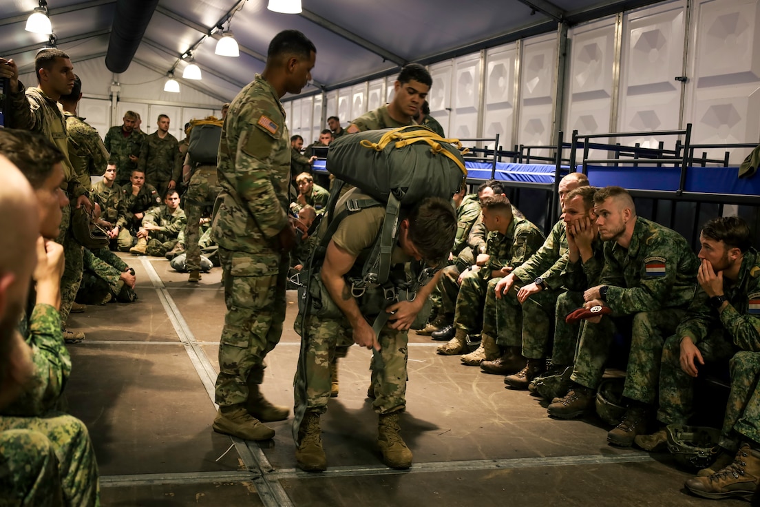 Chief Warrant Officer 2 Joshua Martinez and Chief Warrant Officer 2 Carlos Benitez assist one another in adjusting the parachute rig worn by Staff Sgt. Tristyn Kennedy during a Jumpmaster Personnel Inspection demonstration at Jump week Kamp, Netherlands.