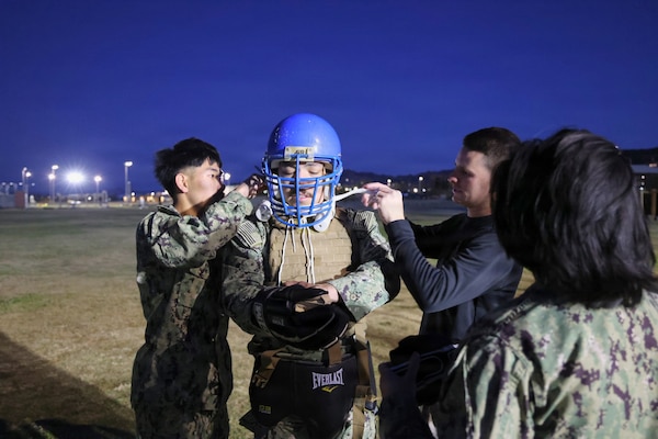Master-at-Arms 3rd Class Thuan Nguyen (left) and Staff Sgt. Joshua Hutchinson (right) assist Religious Program Specialist 2nd Class Diego Escalante with putting on his gear before a pugil sticks match during a Marine Corps Martial Arts Program Tan Belt course at Marine Corps Air Ground Combat Center Twentynine Palms on Dec. 15, 2025. The training bolsters interoperability with Marines to provide war-ready medical care, increases lethality and reinforces the warrior ethos. (U.S. Navy photo by Christopher Jones, Navy Medicine Readiness and Training Command Twentynine Palms / Naval Hospital Twentynine Palms public affairs officer).