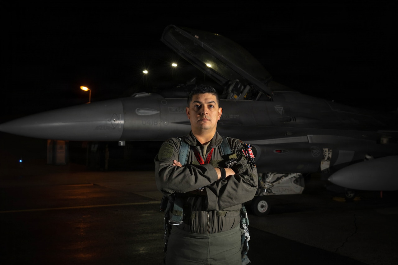 A man in a flight suit stands with his arms crossed in front of a large military fighter jet.