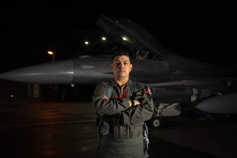 A man in a flight suit stands with his arms crossed in front of a large military fighter jet.