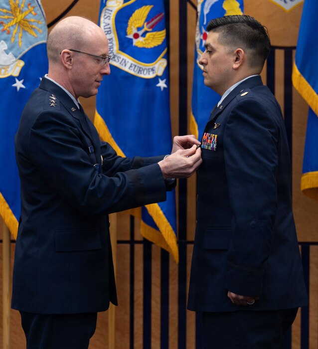 A man in a military dress uniform pins a medal to another man in similar attire as they stand in front of a wall of flags.