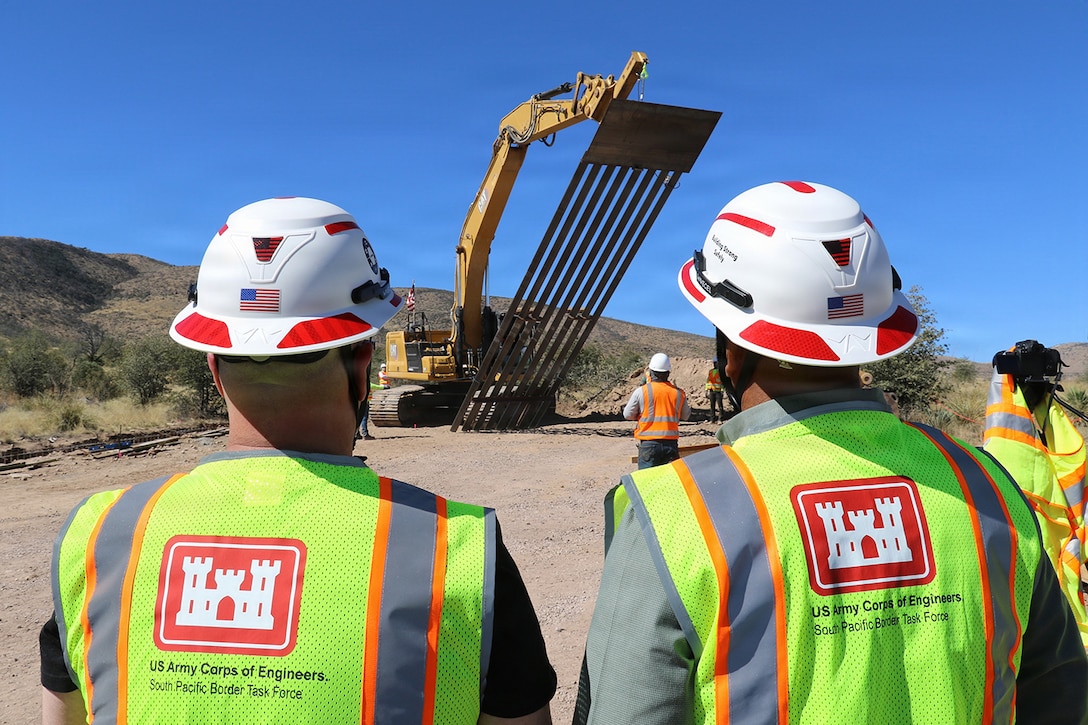 A construction vehicle lifts a large brown bridge section as works in hard hats watch.