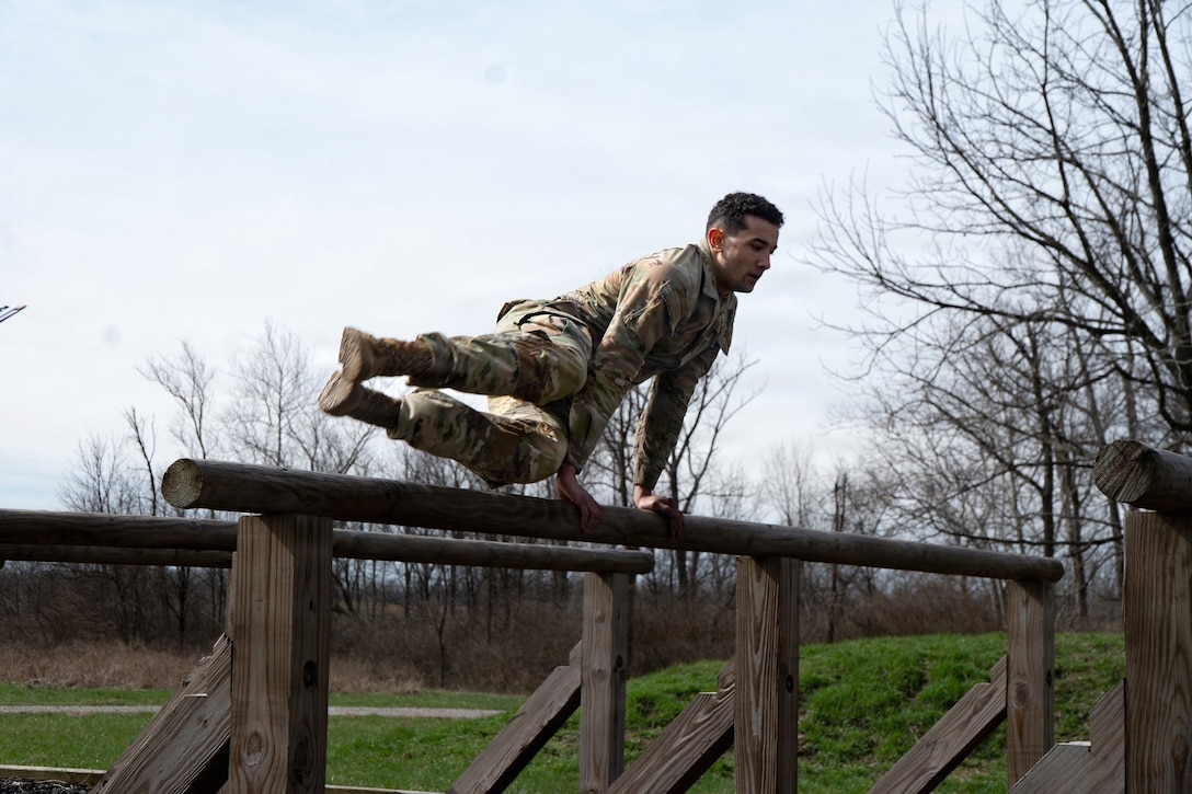 A soldier jumps over a wooden obstacle course in a field under a bright sky.