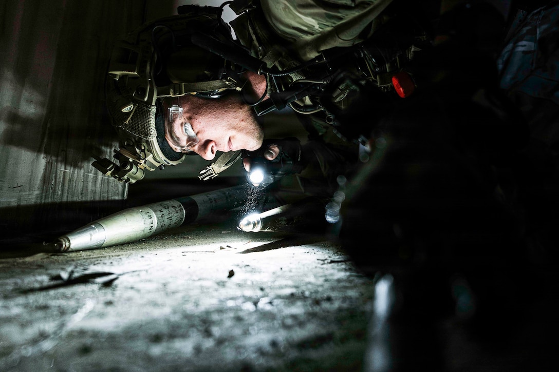 An explosive ordnance disposal technician wearing a helmet and protective eyewear lies on the ground while using a flashlight to inspect ordnance in the dark.