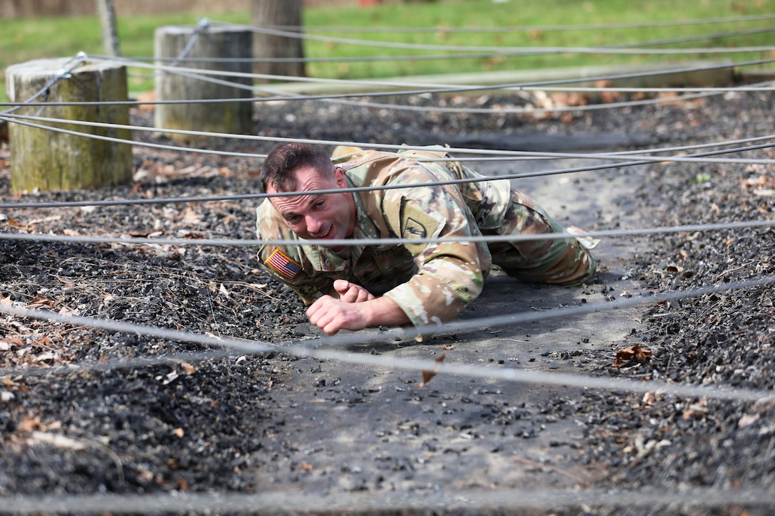 A soldier crawls on pavement lined by black rocks under barned wire during the day.