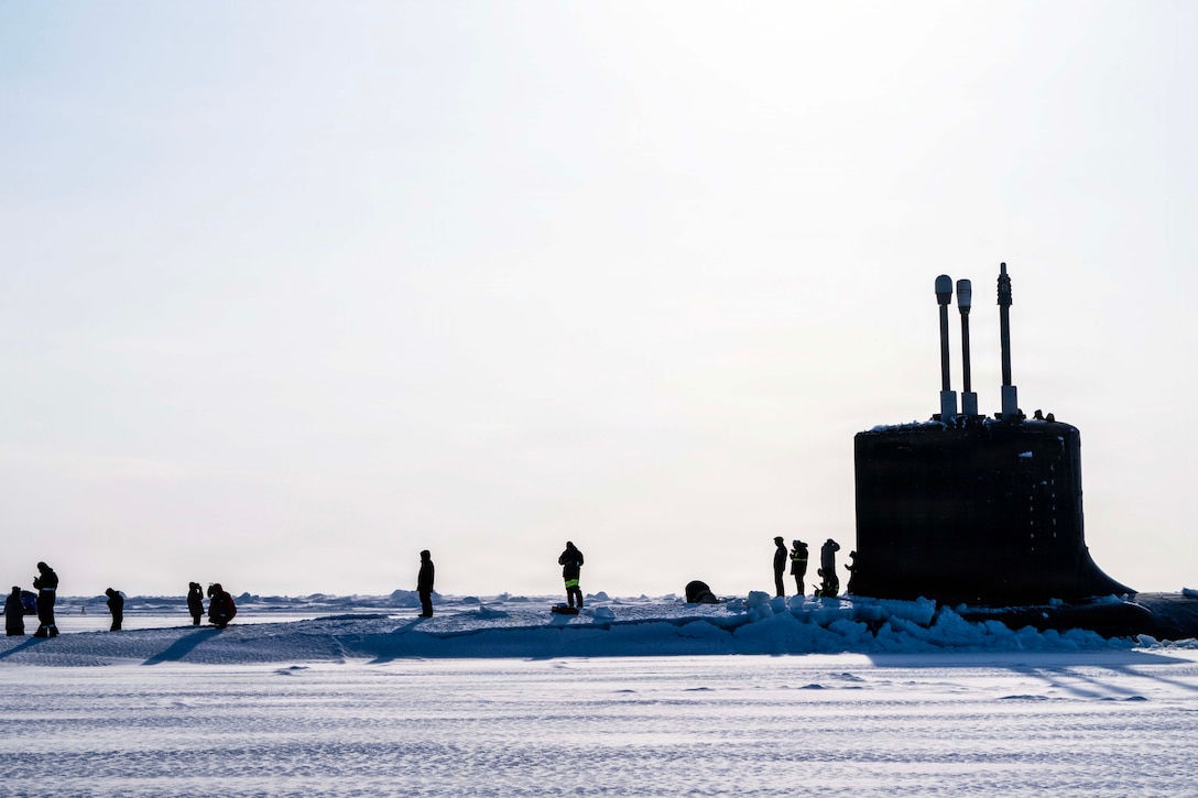 Sailors stand on a snowy submarine surrounded by a snowy, icy surface under a bright sky.