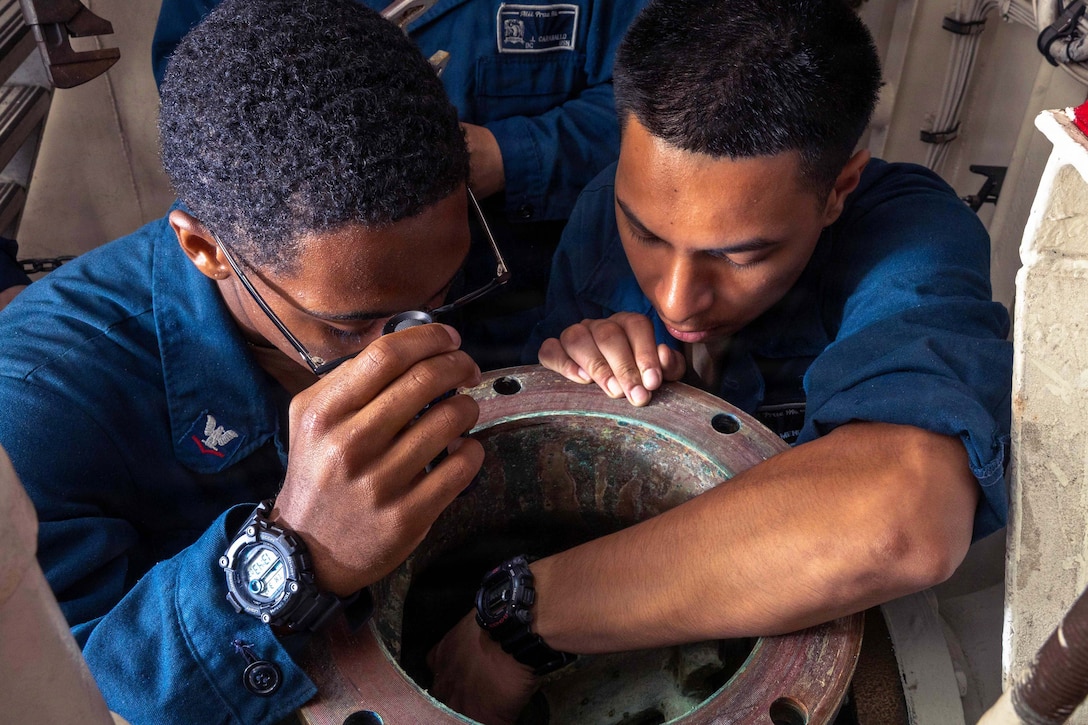 A close-up of two sailors in blue jumpsuits inspecting an inductor.