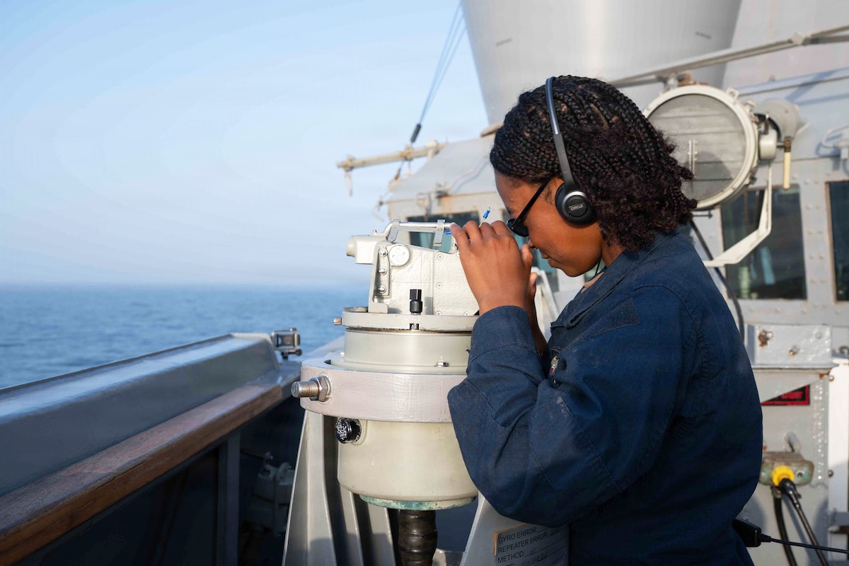 A sailor wearing a headset uses an alidade on the side of a ship at sea during the day.