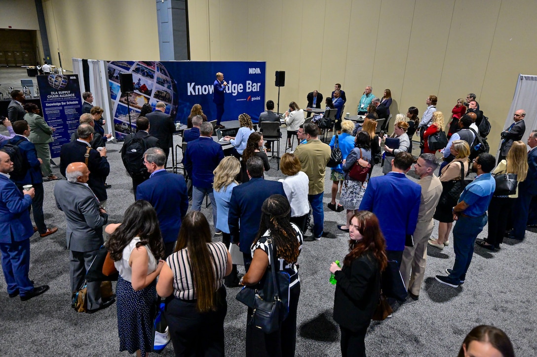 A group of people in business attire stand in a exhibit hall listening to a man in a blue suit speak.