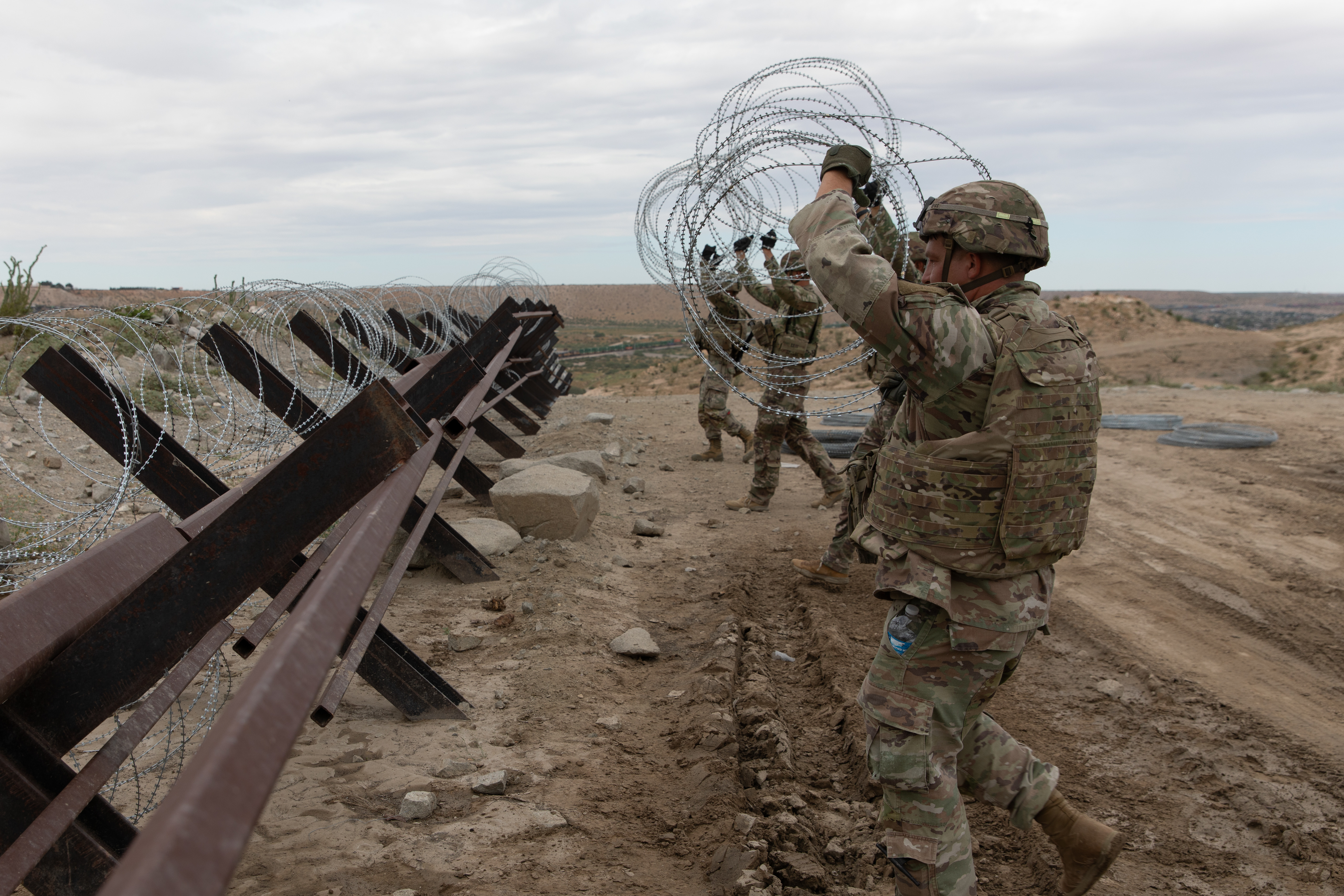 People wearing military camouflage uniforms move concertina wire over a barrier fence in desert terrain.