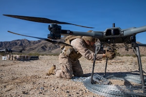 A man wearing a camouflage military uniform works on a drone in mountainous desert terrain.
