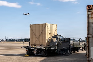 A K-loader with heavy cargo on it sits on the flightline as aircraft fly overhead.