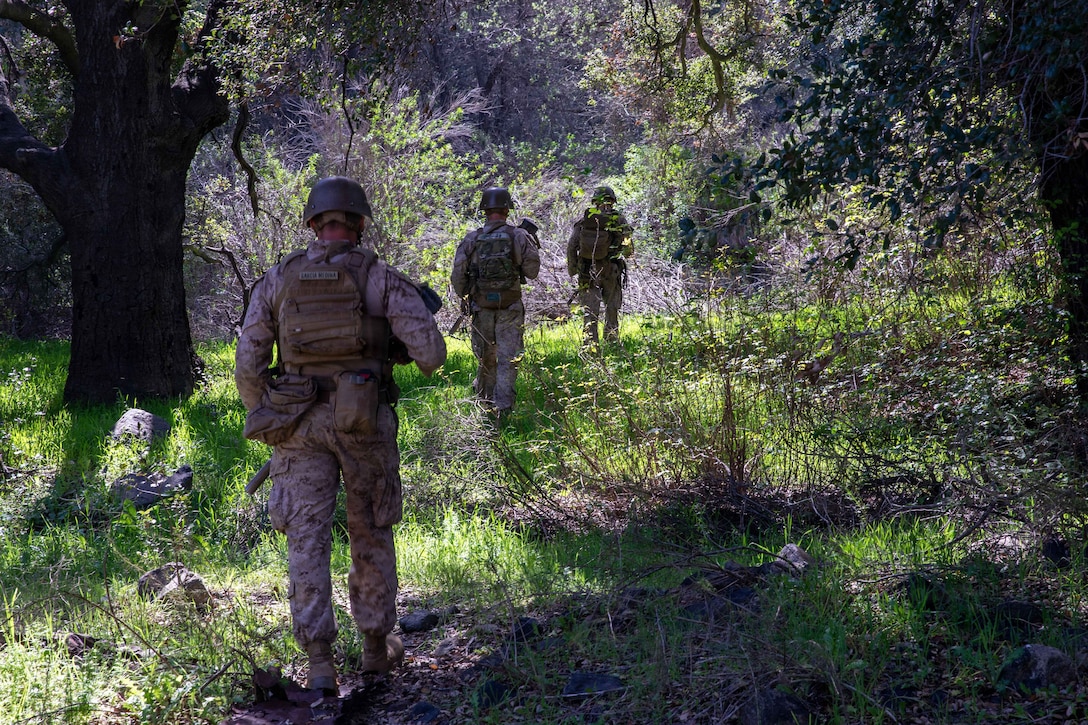A line of three Marines walk along a wooded area in dappled sunlight.
