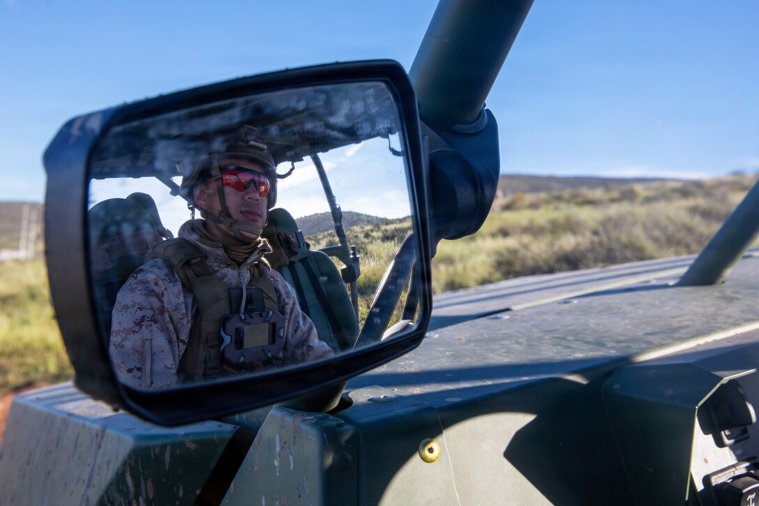A Marine is reflected in the mirror of a vehicle that's facing a wide,  grassy area on a sunny day.