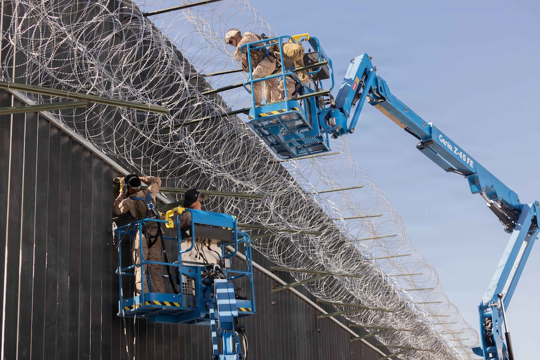 Two Marines in elevated truck ladders work on a tall fence with concertina wire at the top.