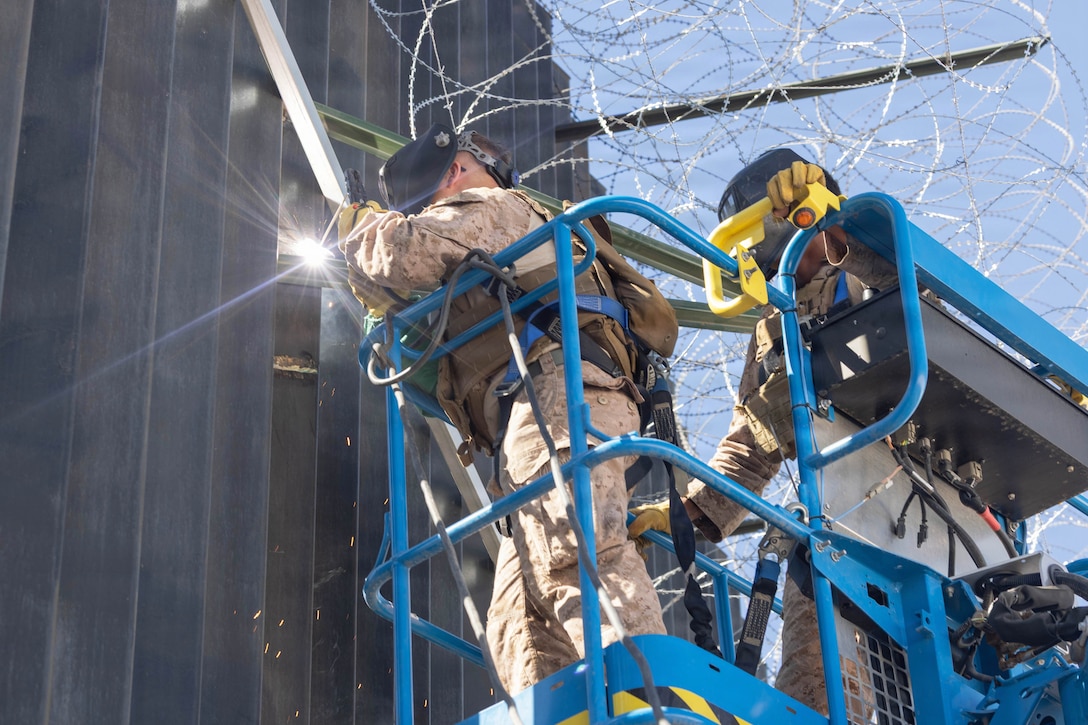 Two Marines in a raised ladder and wearing welding gear work on a large fence with concertina wire overhead.