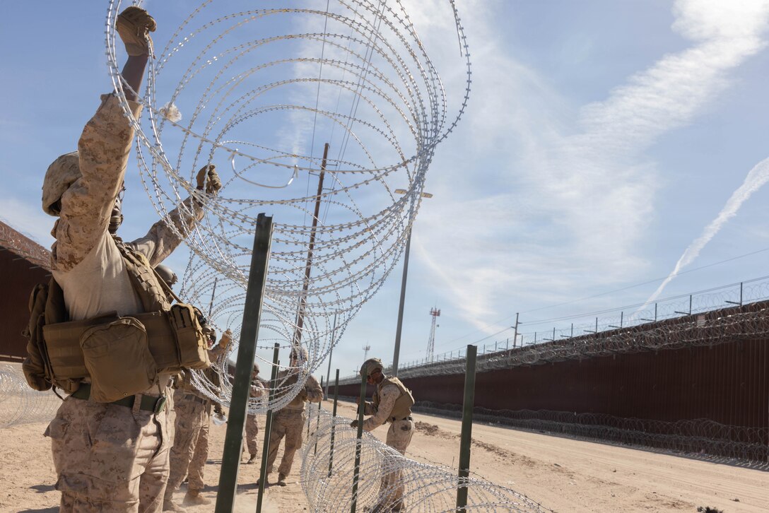 A small group of Marines on the ground arrange concertina wire overhead along a fence line in a dry desert-line area.