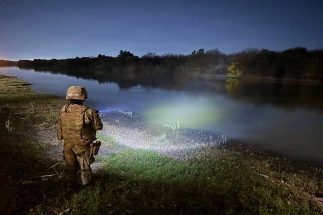 A soldier shines a light across a river at night, with trees lining the other bank.