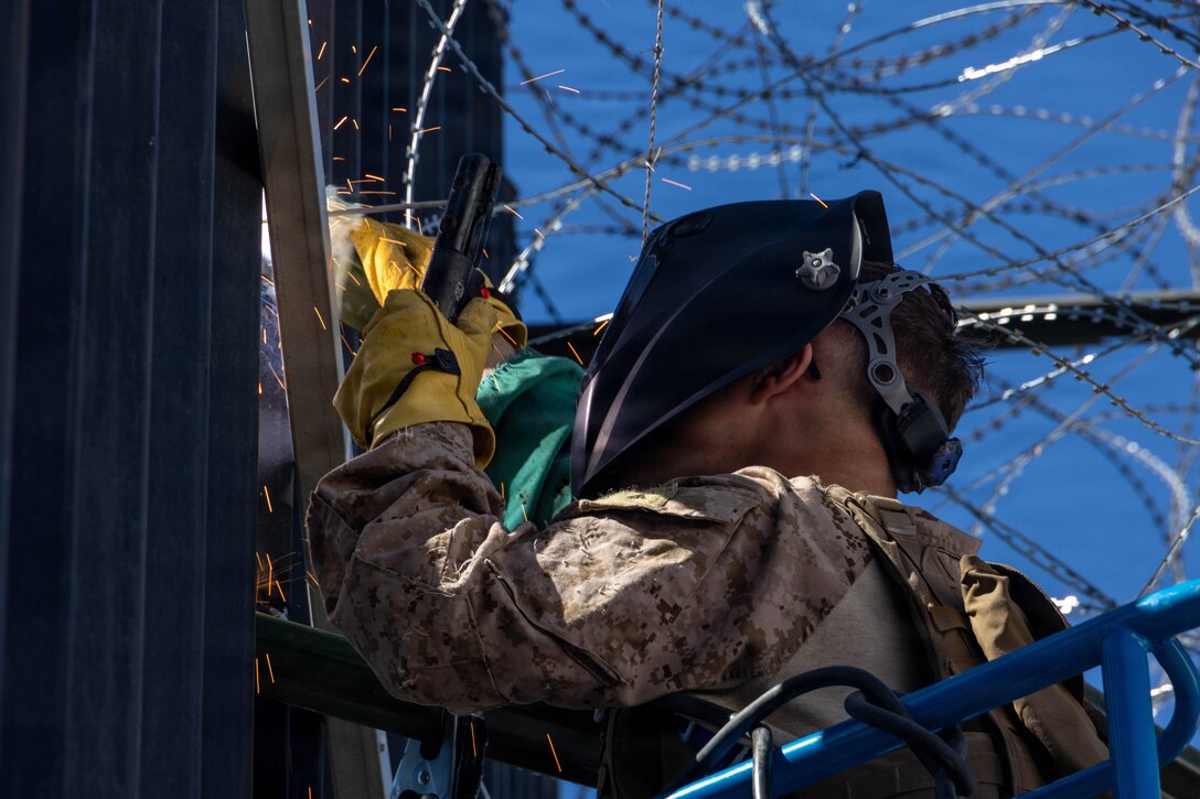A Marine in welding gear works on a section of the southern barrier with concertina wire overhead.