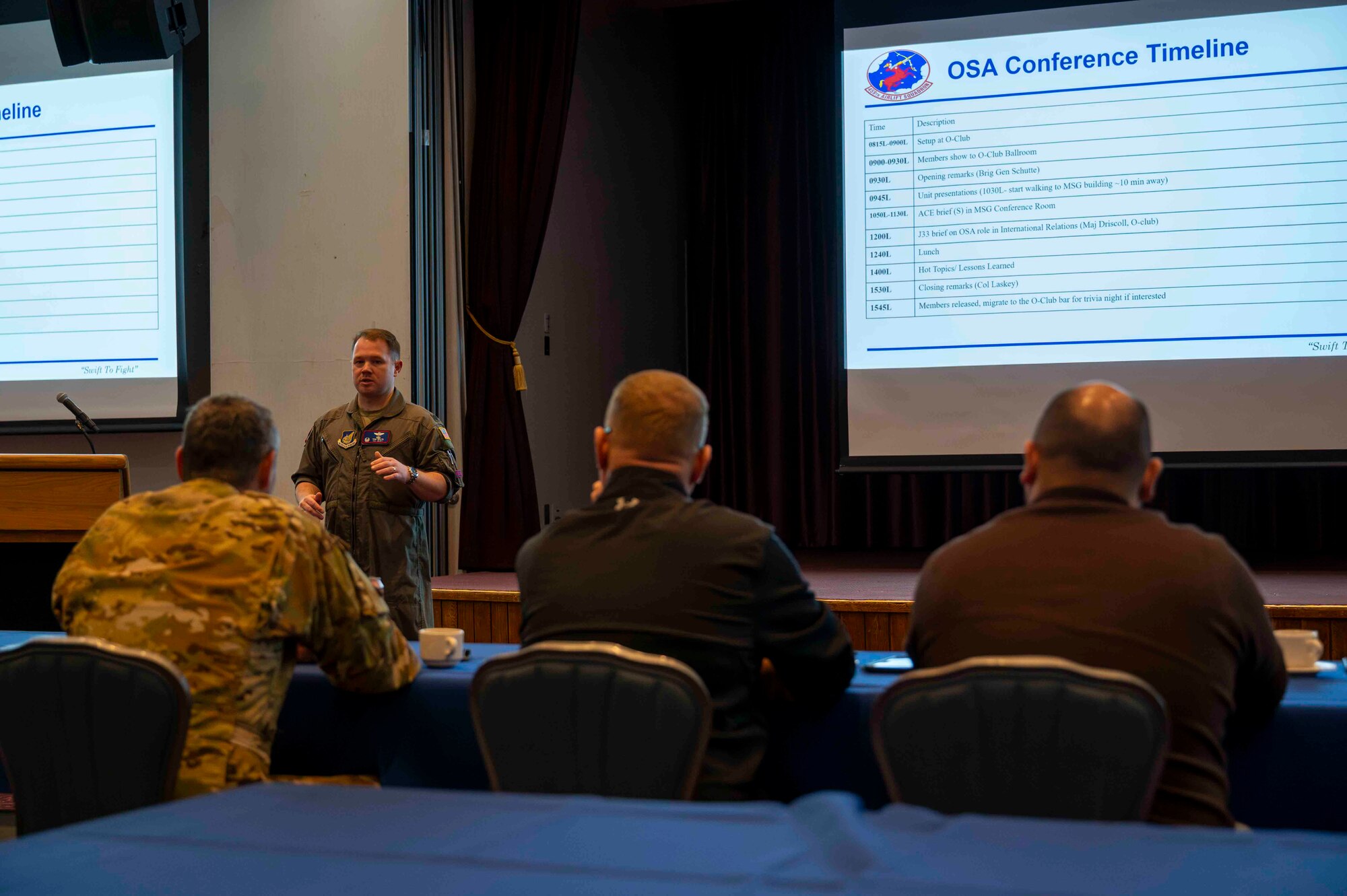 A military member speaks in front of three members sitting in chairs.