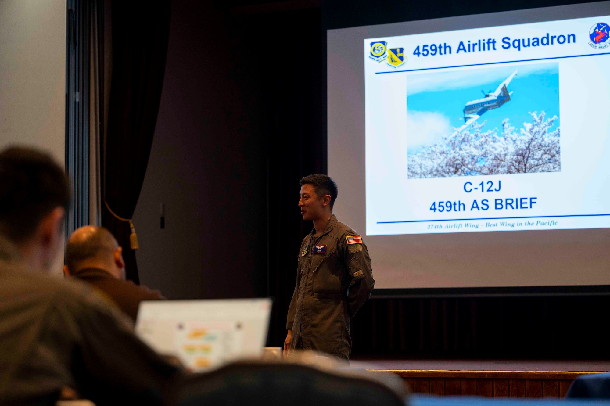 A military member stands in front of a projector showing a C-12J Huron.