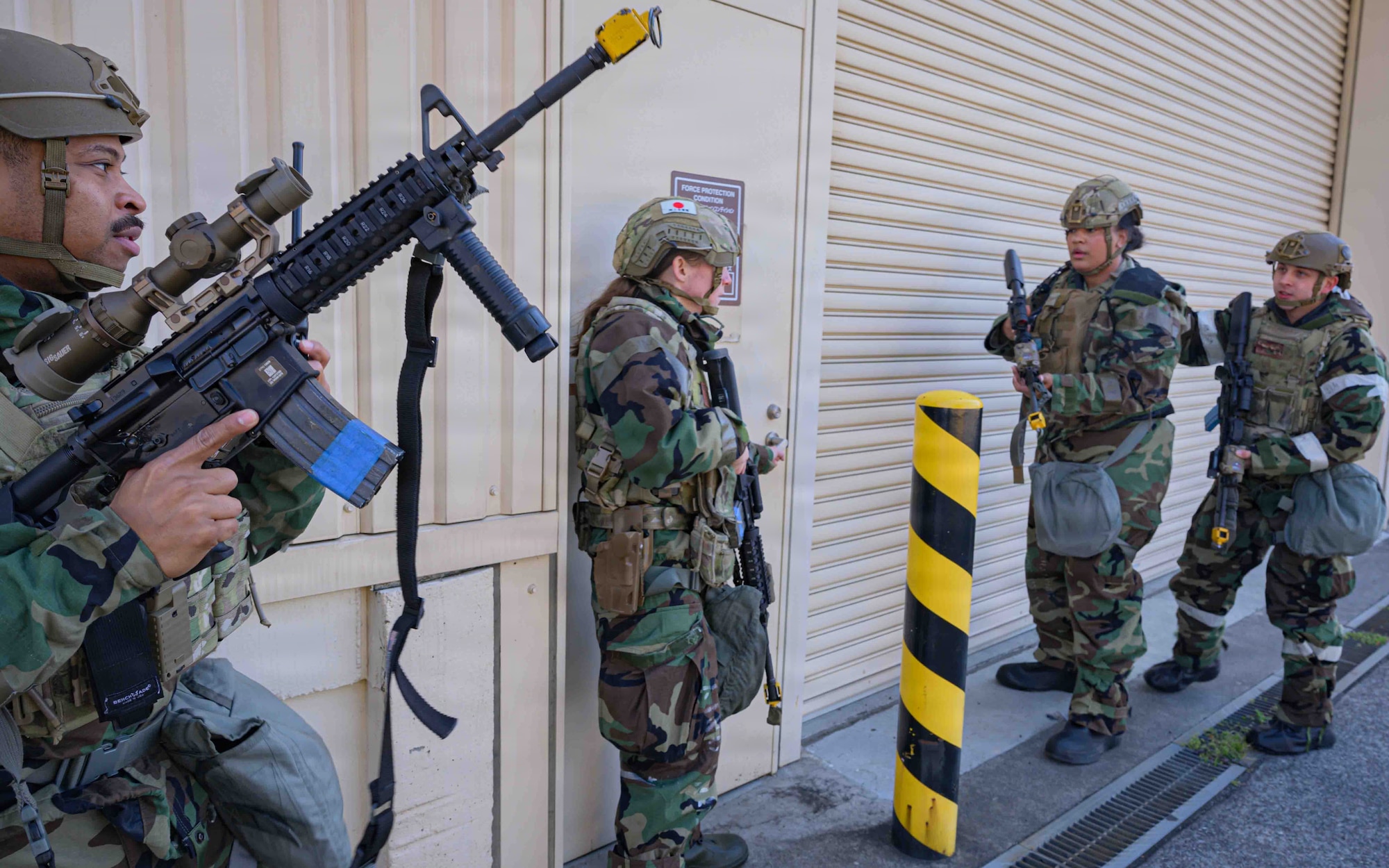 Four Airman with serious faces gather around a tan door while holding onto their black rifles. They are wearing green and black camouflage coats and tan helmets.