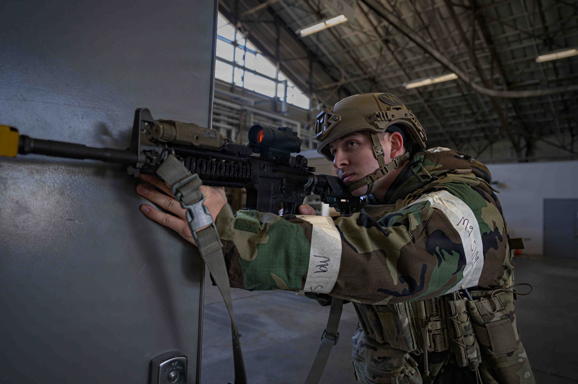 An Airman with a serious face aims his black rifle while holding open a grey door. He is wearing a green and black camouflage coat and a tan helmet.