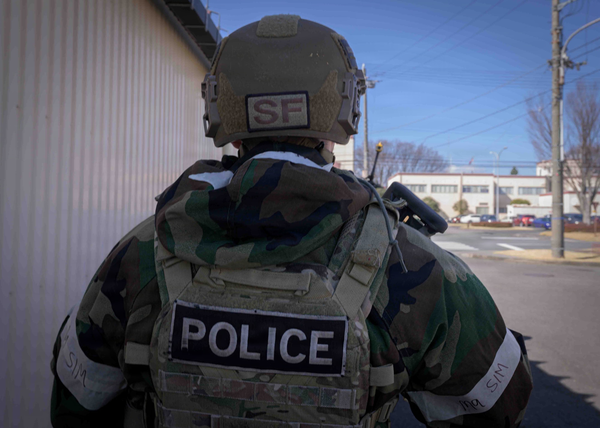 The view of the back of an Airman looking out towards a road. He is wearing a black and green camouflage pattern coat with a helmet that has a brown patch with the letters SF and a weighted vest that has POLICE written on it in black and white.
