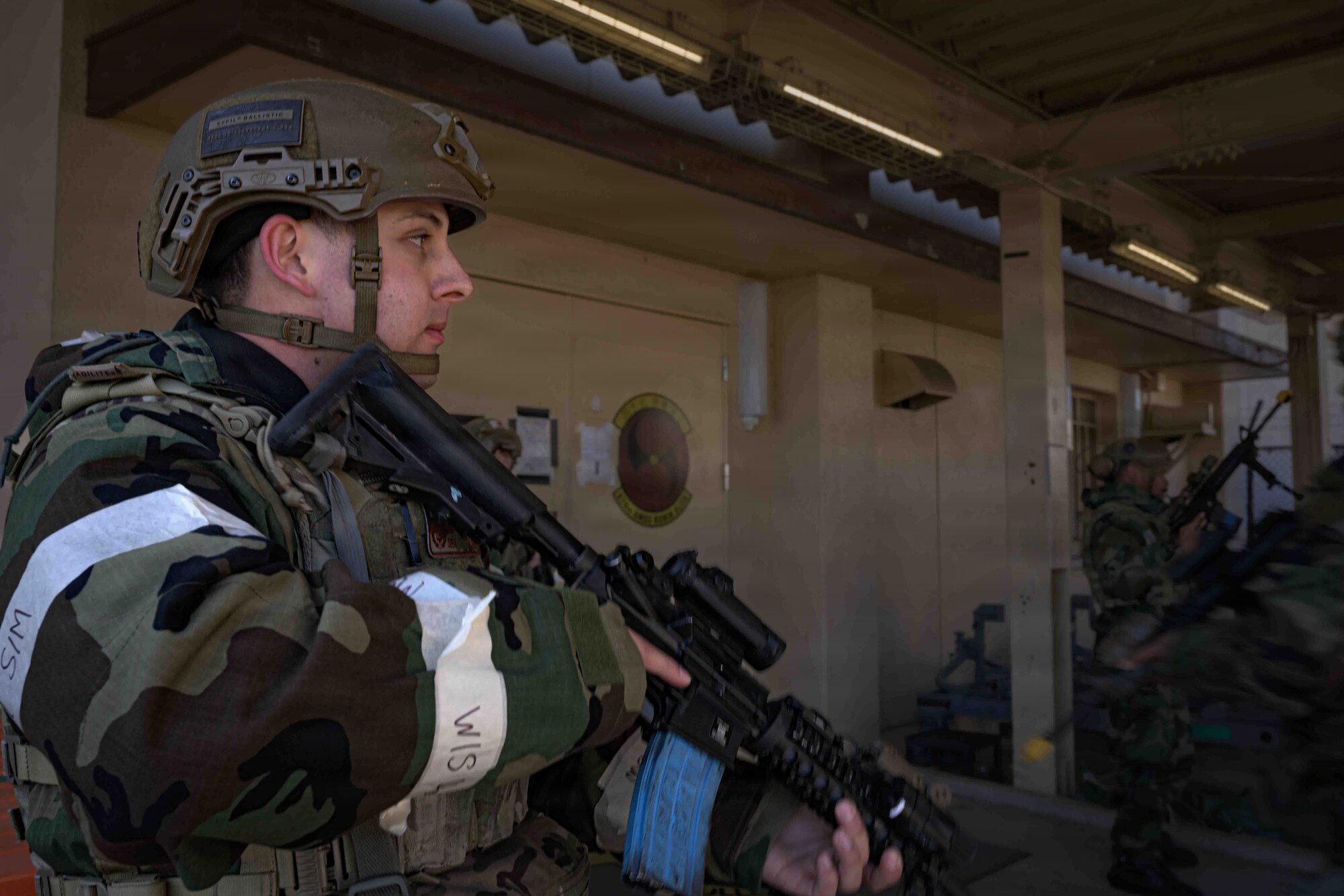 An Airman with a camouflage green and black protective coat holds a black rifle aiming down while looking forward. He wears a tan helmet that exposes his serous but calm expression on his face.
